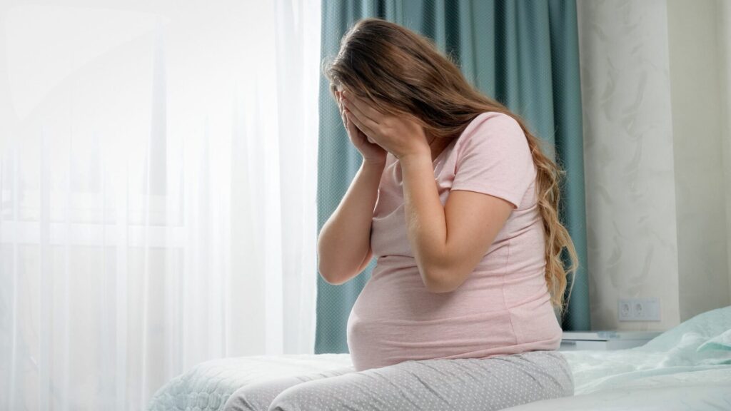Sad crying pregnant woman suffering from depression sitting on bed and holding her head. Concept of