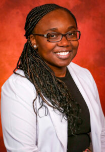 A smiling woman with braided hair, wearing glasses and a white lab coat, against a vibrant orange background.